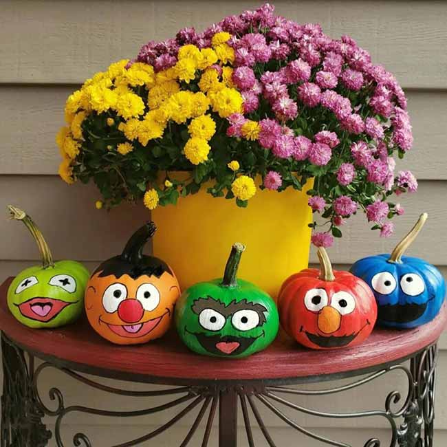 Five small pumpkins are cleverly painted to look like the friendly faces of Sesame Street characters, sitting on a dark red table below a vibrant pot of yellow and purple flowers.