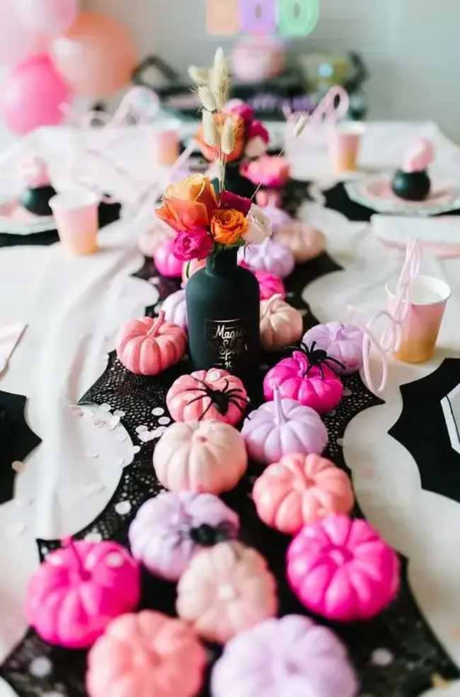 A long party table features a spooky black bat-shaped runner that is covered in a cascade of mini pumpkins painted in shades of hot pink, light pink, and purple.