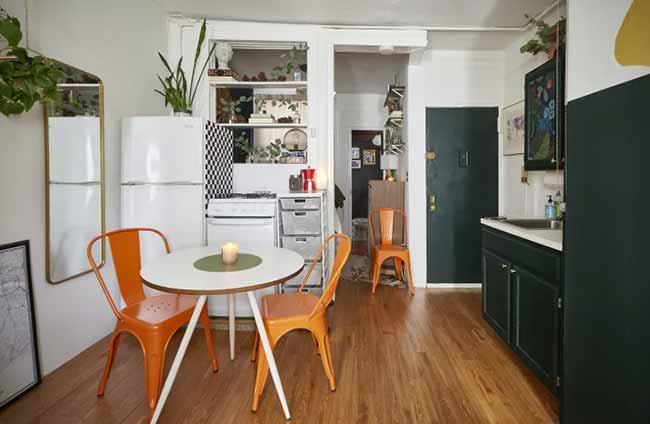 In this small living room with dining table, bright orange chairs pop against the white-and-black checkered kitchen and dark green cabinets.