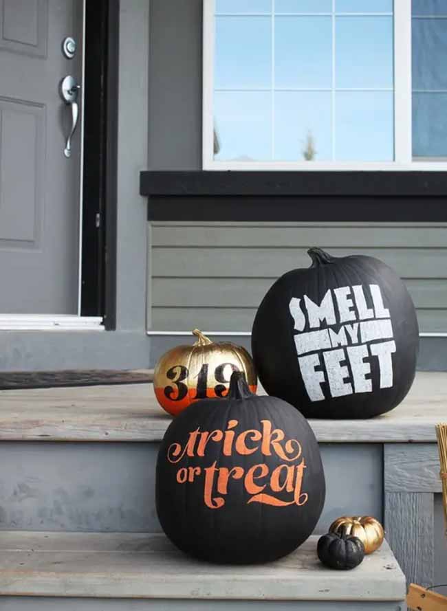 Three pumpkins with modern lettering sit on a porch step; one says "Trick or Treat" in fancy orange script while another cheekily says "Smell My Feet" in bold white letters.