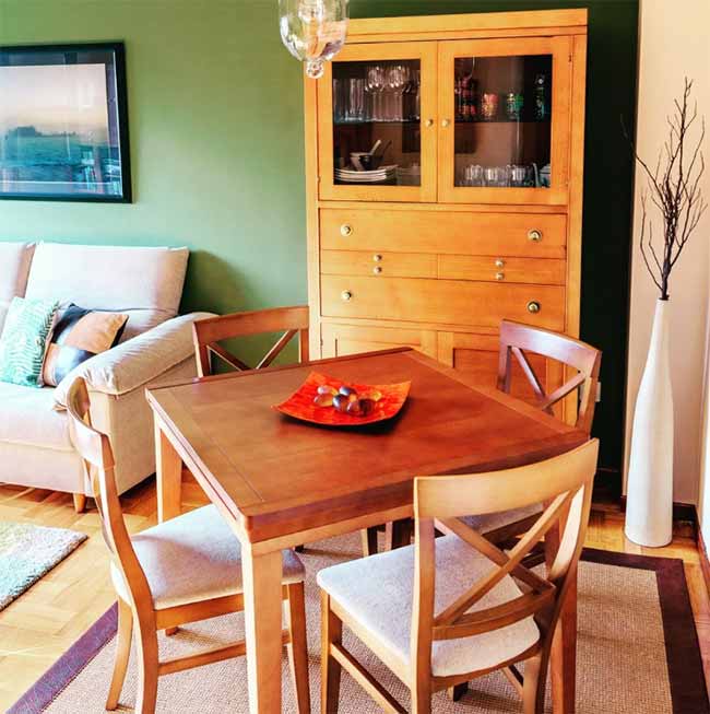 A square wooden table with upholstered chairs sits against a two-tone green wall, anchored by a classic wood hutch for charm.