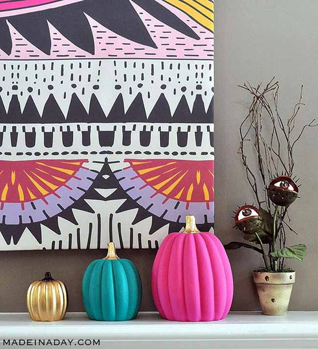 Three small, sleek pumpkins painted in solid gold, teal, and hot pink sit on a shelf next to a spooky plant with eyeballs and a colorful geometric art print.