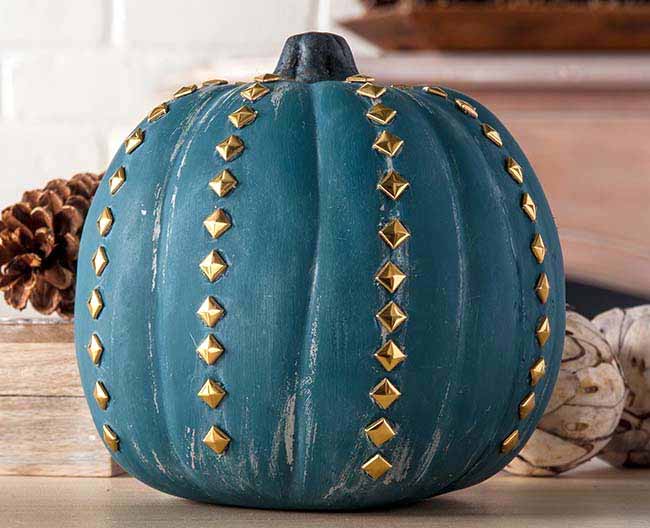 A pumpkin painted a dusty teal color is decorated with vertical lines of gold pyramid studs, giving it a chic and edgy vibe.