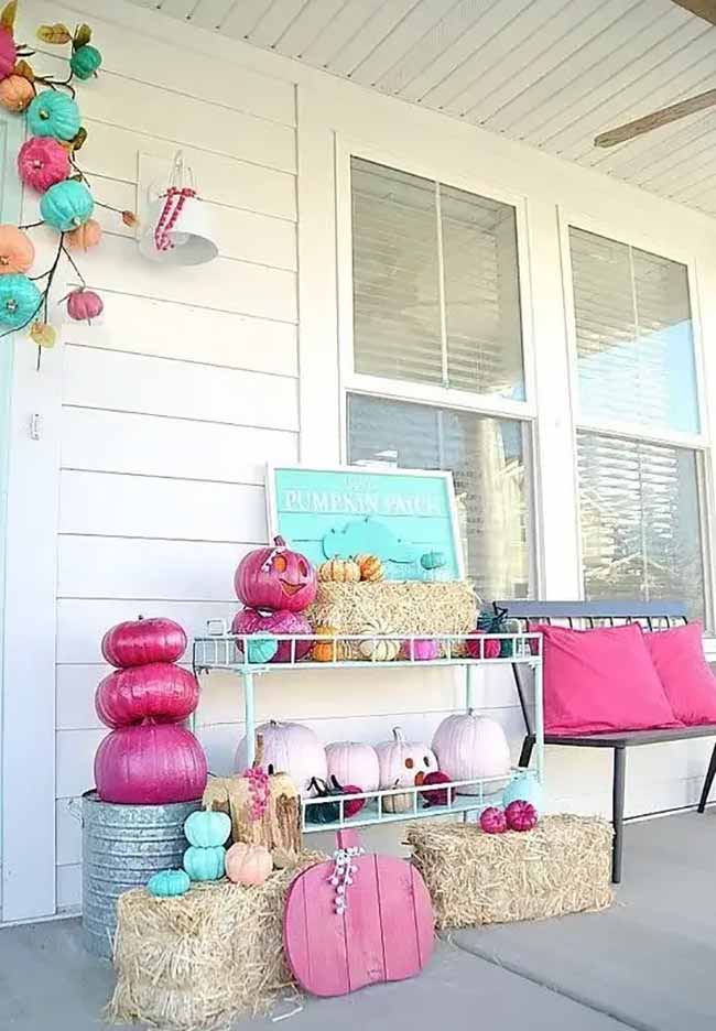 A front porch is decorated with a cheerful and unconventional pink Halloween theme, featuring hay bales, a shelf full of pink-painted pumpkins, and a matching pink pumpkin garland.