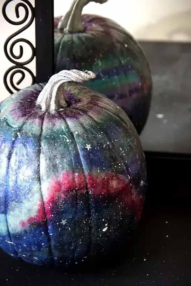 Two pumpkins are painted with a deep, cosmic galaxy design, featuring swirls of purple, blue, and red, and are lightly dusted with white paint and tiny star glitters.