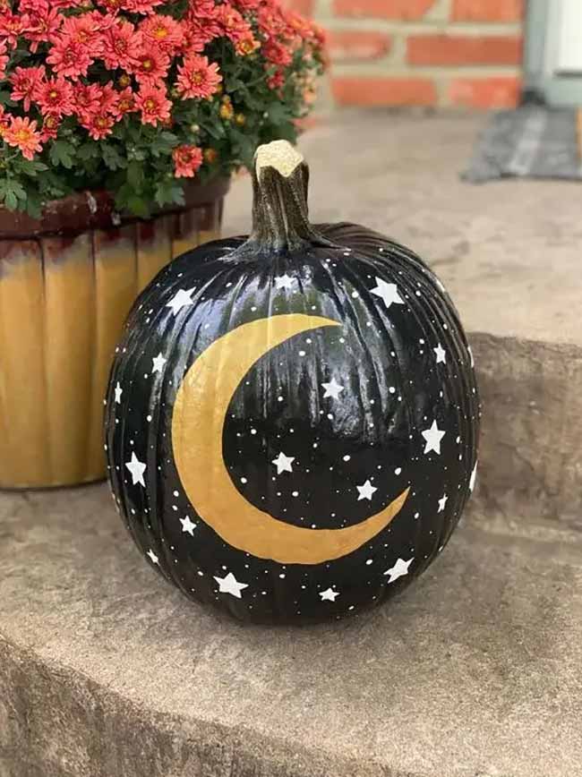 A glossy black pumpkin is painted like the night sky, with a golden crescent moon and scattered white stars.