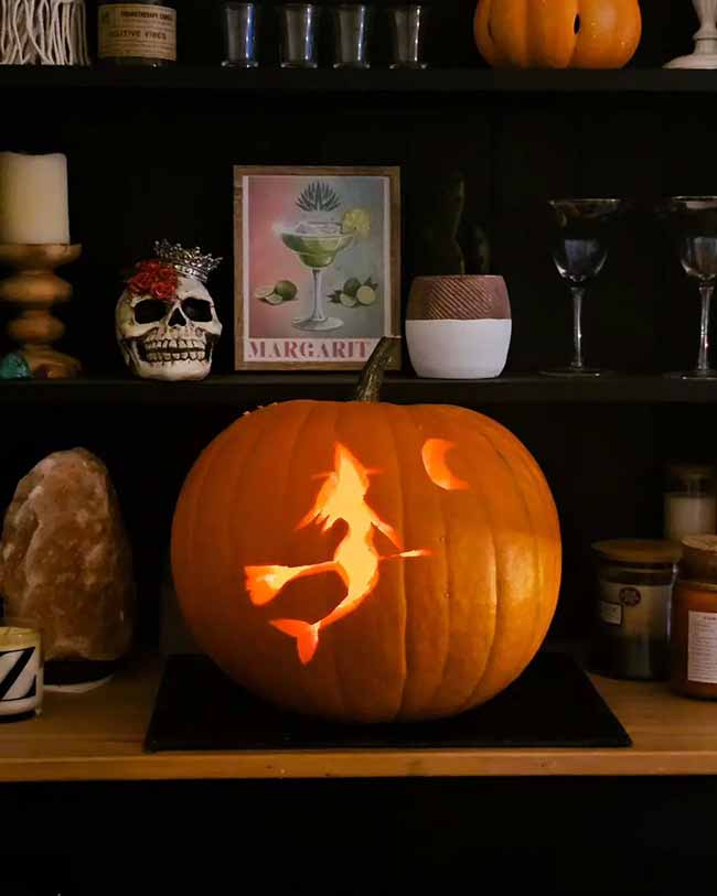 Placed on a decorated shelf, this jack-o'-lantern features a classic silhouette of a witch flying on her broomstick.