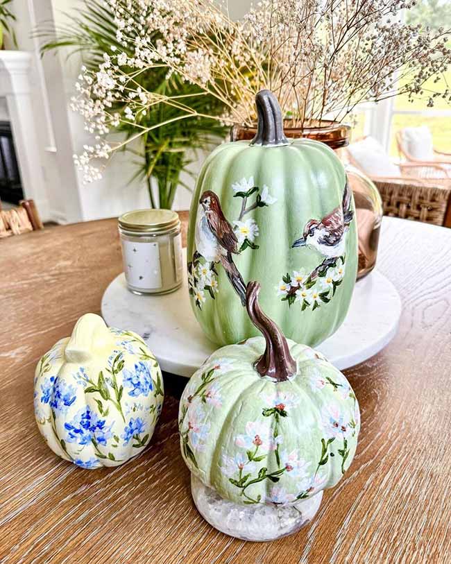 Three pumpkins are elegantly painted with delicate floral patterns; the largest sage green one also features two small birds perched on branches, making a lovely fall centerpiece.