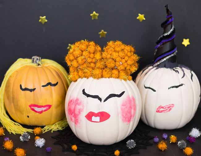 Three pumpkins are transformed into playful characters with painted-on makeup and fun hair, including one with yellow yarn pigtails, another with orange pom-pom space buns, and a third with a sparkly witch horn.