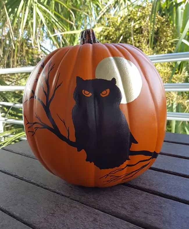 An orange pumpkin is painted with a silhouette of a black owl with intimidating orange eyes, perched on a branch in front of a big, golden full moon.