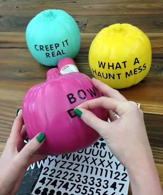 A bright pink pumpkin with black letter stickers sits in front of yellow and teal pumpkins painted with funny phrases like “Creep it real.”