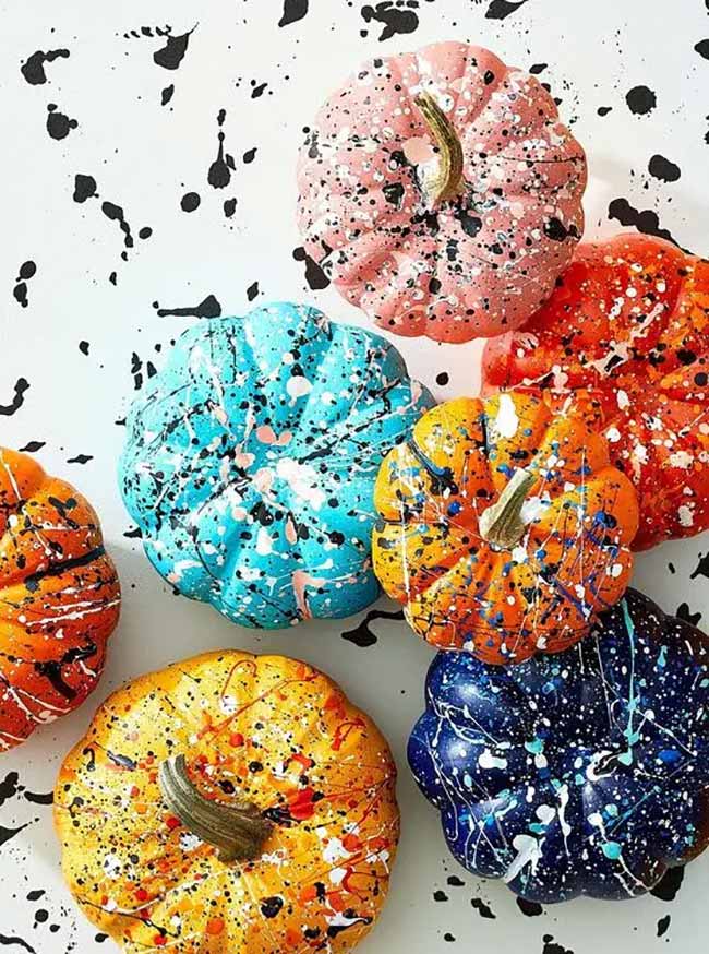 A variety of colorful pumpkins in blue, pink, orange, and yellow are laid out on a white surface, all decorated with an splatter of black and white paint.