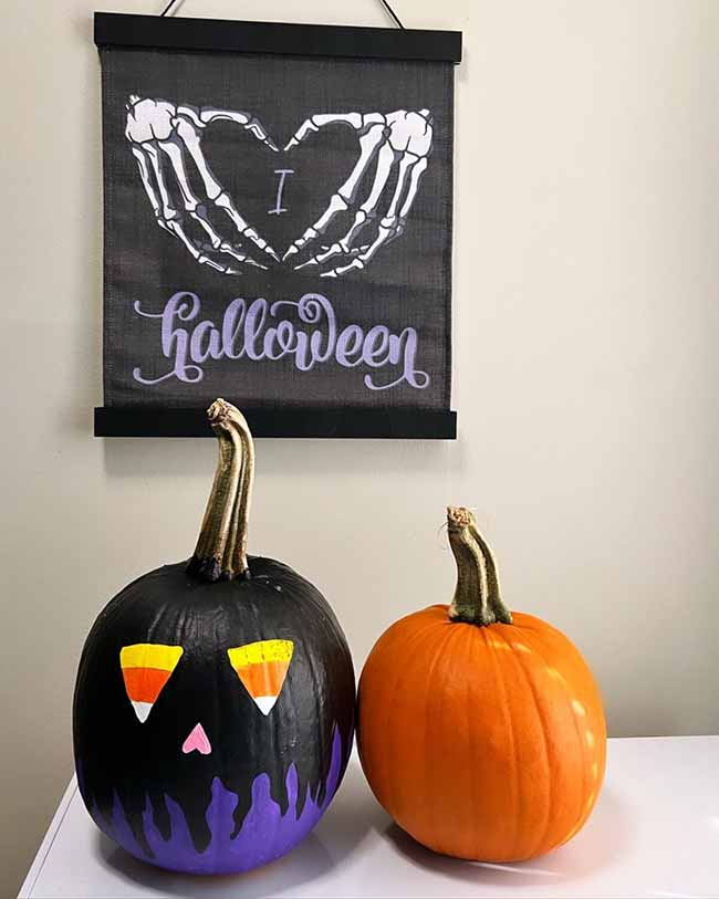 A black pumpkin is painted with a spooky monster face, its eyes drawn to look like candy corns, with purple flames dripping from the bottom, beside a plain orange pumpkin.