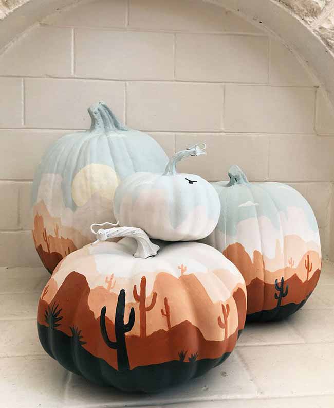 A group of pumpkins is painted to create one continuous desert scene at dusk, showing rolling brown hills and tall saguaro cacti against a soft, pale blue sky.