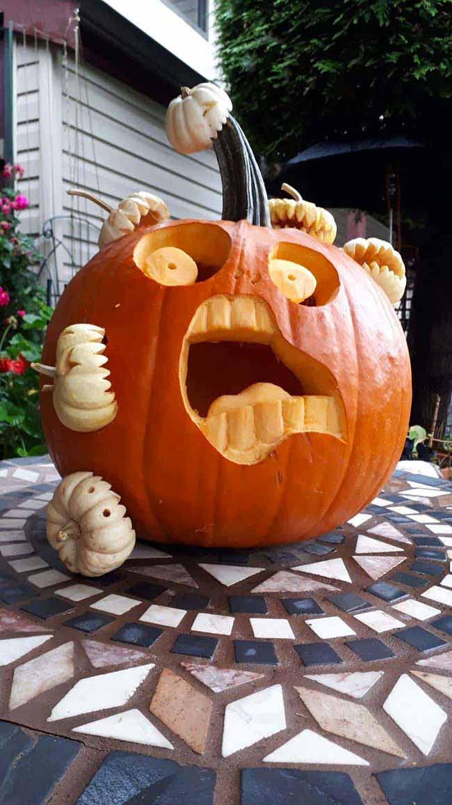 A giant pumpkin with a wide-open, screaming mouth sits on a table, with several smaller pumpkins stuck to it like they're being eaten. It's a really creative pumpkin carving idea.