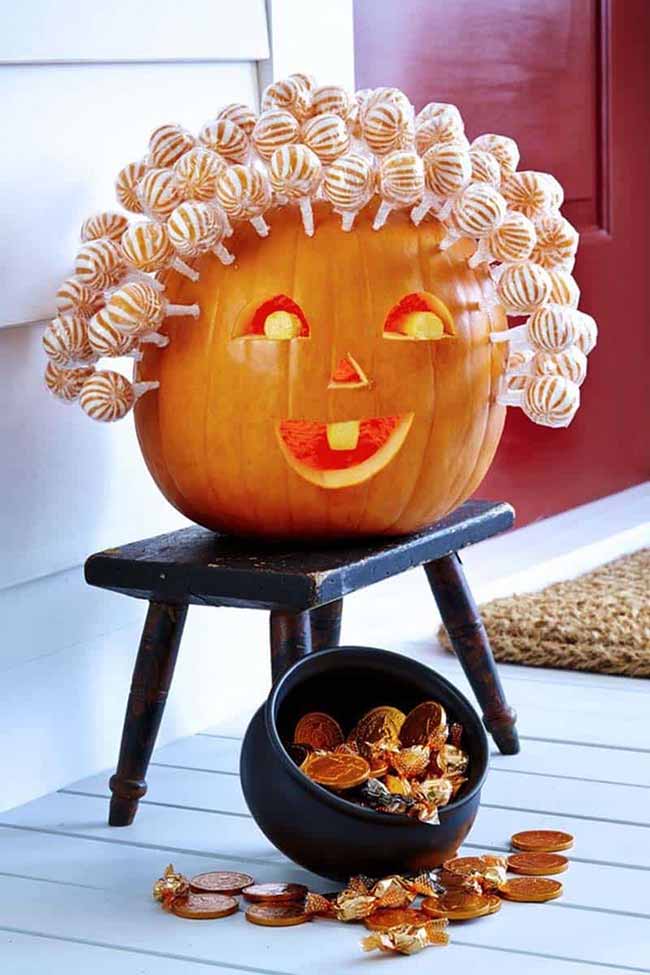 This pumpkin is sitting on a small stool, with a happy face carved into it and a head full of colorful lollipops. Next to it is a bowl filled with candy, ready for trick-or-treaters.