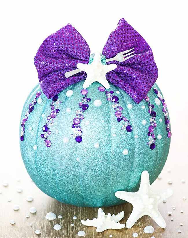 A teal glitter pumpkin inspired by “The Little Mermaid,” decorated with cascading purple and white rhinestones, a sparkly purple bow, and a tiny white fork tucked into the ribbon.