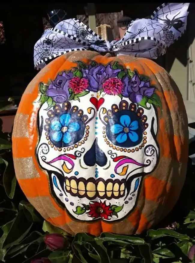 An orange pumpkin features a vivid sugar skull design, with floral eyes and a crown of purple roses, painted in the classic Dia de los Muertos style.