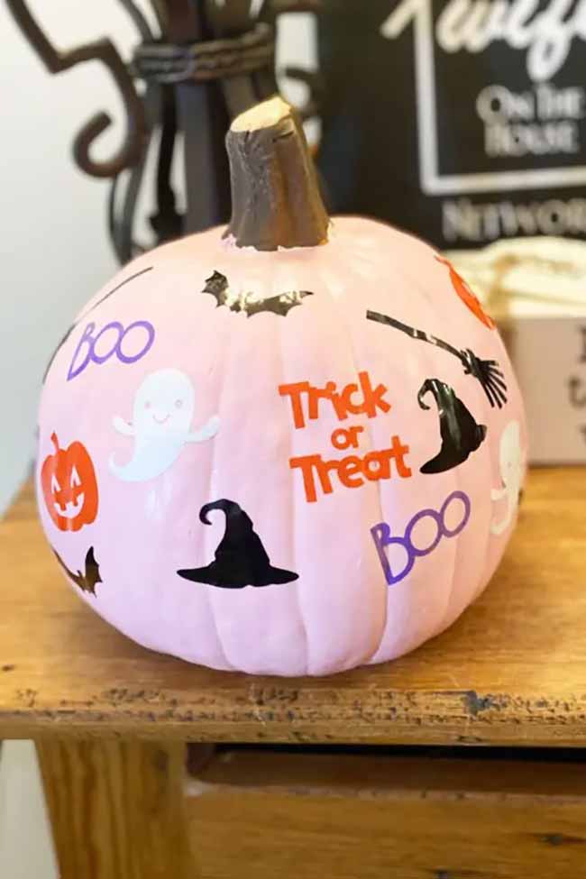 A pastel pink pumpkin is covered in playful Halloween stickers, including ghosts, bats, witch hats, and the words “Trick or Treat” in bright orange letters.