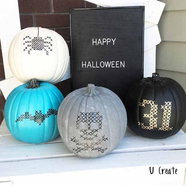 Four pumpkins are painted in a cross-stitch style: white with a spider, blue with bats, gray with a skull, and black with a golden “31.”