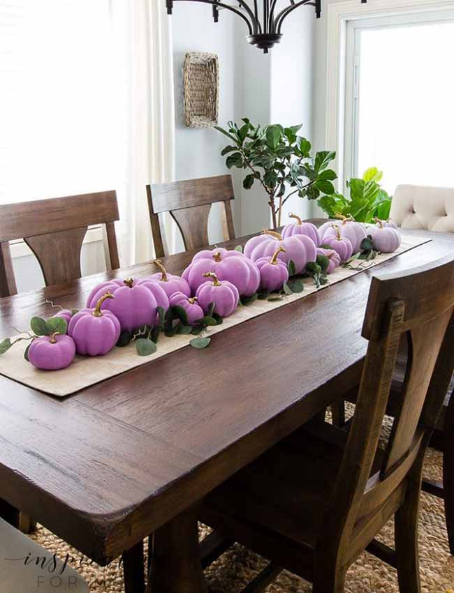 A dining table centerpiece features pumpkins in mixed sizes, painted lavender and arranged with green leaves for a soft, elegant look.