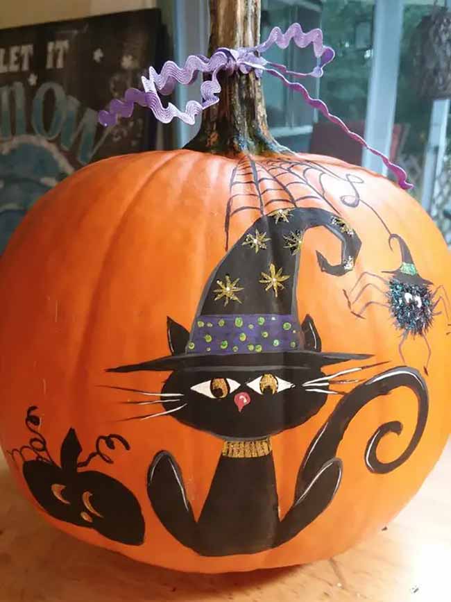 A large orange pumpkin is painted with a black cat in a starry witch hat, accompanied by a glittery, googly-eyed spider hanging from a web.