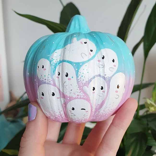 A small pumpkin is covered in a cluster of adorable little white ghosts floating over a pastel pink and blue background.