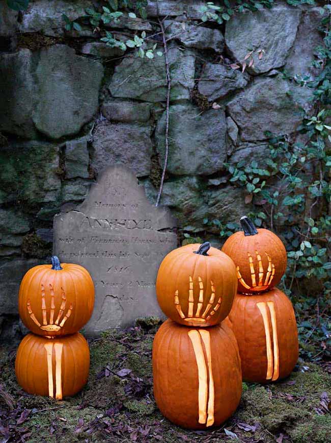 Placed in a spooky graveyard, two stacks of pumpkins have carved hands reaching out. The glowing light from inside makes the hands look skeletal.