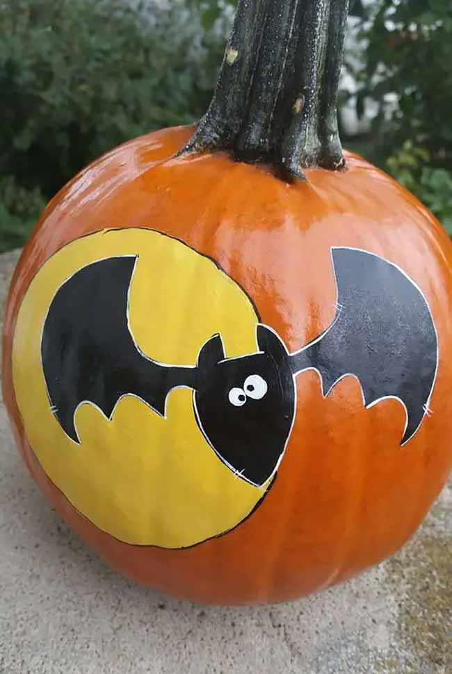 An orange pumpkin is set against leafy green foliage and features a stylized black bat flying in front of a bright yellow moon.