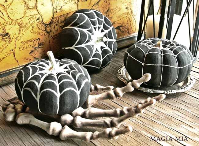 Two black pumpkins display intricate white spiderweb designs, one on a silver dish and the other on a white skeleton hand for a creepy table centerpiece.