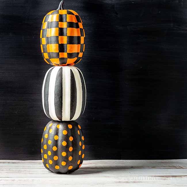 A stack of three whimsical pumpkins sits against a dark background, each painted with a different black-and-white pattern: checkerboard on top, bold vertical stripes in the middle, and polka dots at the base.