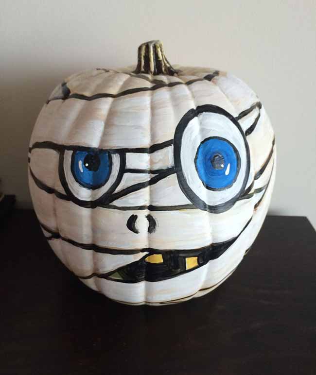 A hand-painted pumpkin resembles a friendly mummy, with thick white lines and wide blue eyes peeking through the bandages.