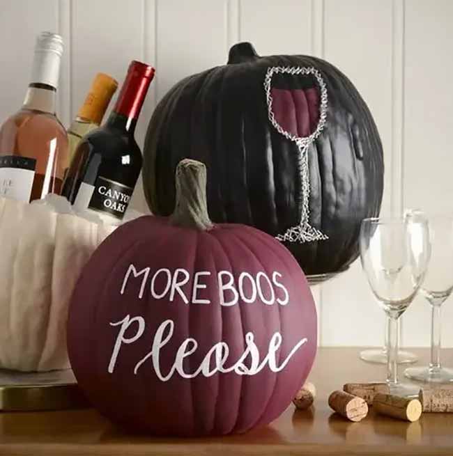 A dark reddish-purple pumpkin is painted with the playful phrase “More Boos Please” in white script, perfect for Halloween décor.