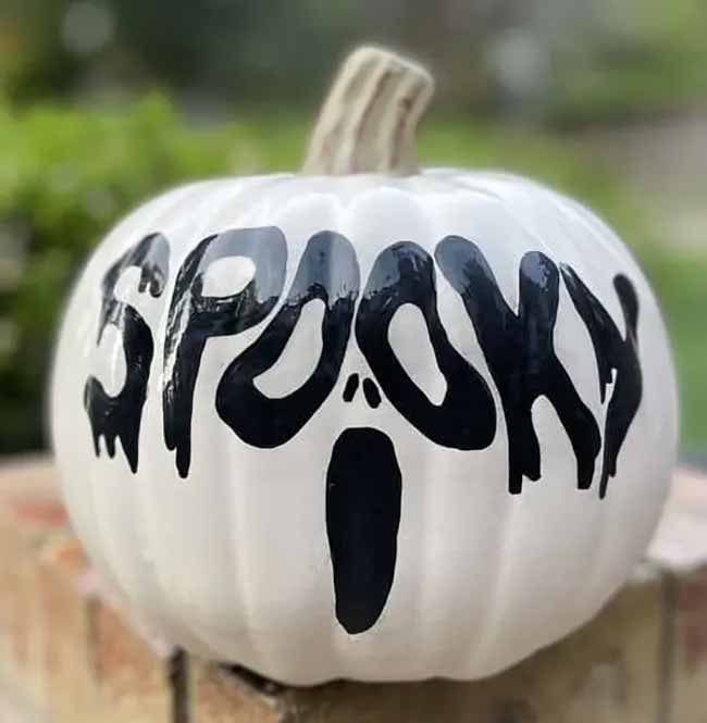 A stark white pumpkin is painted with the word “SPOOKY” in drippy black letters above a simple ghost face.
