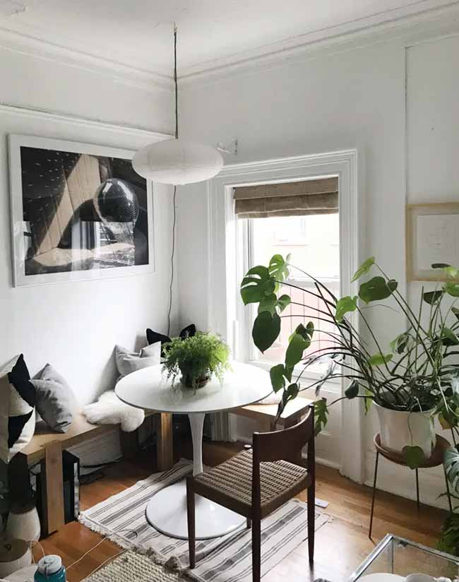 A white tulip table with a wooden bench and wicker chair fills a sunlit nook with light and greenery.