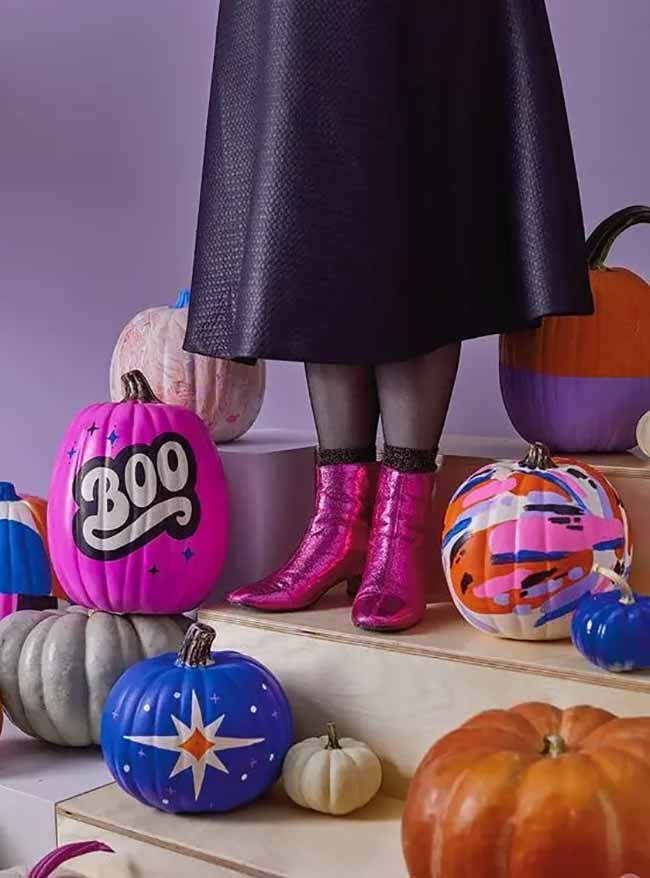 A large bright pink pumpkin painted with the word “BOO” in a retro font sits among colorful pumpkins, alongside a pair of sparkly pink boots.