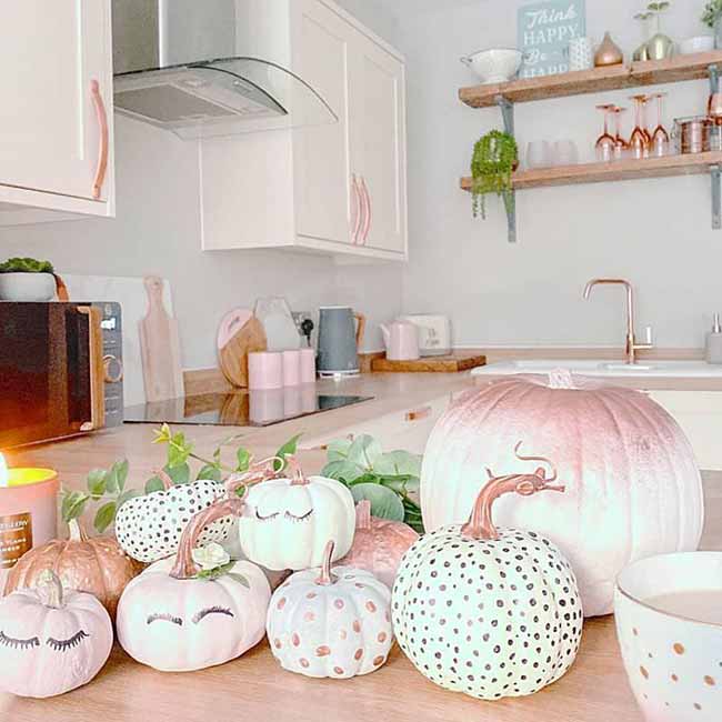 A kitchen counter is filled with soft-toned pumpkins, including pale pinks and whites with dreamy closed eyes and others dotted with shiny metallic polka dots.