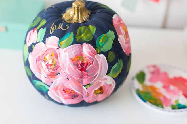 A dark blue pumpkin is adorned with textured pink roses, green leaves, and the word “fall” in gold script near the stem.