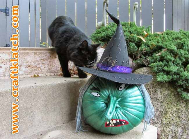 A black cat approaches a pumpkin painted as a witch’s head, complete with a green face, long nose, and tall black hat.