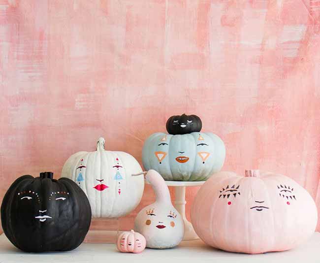 A family of minimalist pumpkins, each painted in soft pastels or black with simple faces, is styled against a pale pink backdrop for a chic, spooky look.