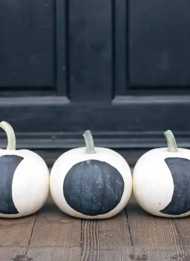 Lined up on a wooden porch, three white pumpkins are simply painted with the different phases of the moon in a matte black color.