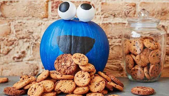This pumpkin is painted a vibrant blue and transformed into a Cookie Monster, with big googly eyes and a black mouth, surrounded by a pile of delicious chocolate chip cookies.