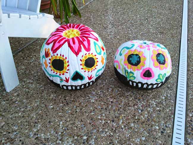 A pair of pumpkins is painted in a colorful Day of the Dead, or Sugar Skull, style, with a flower on top and bright designs around the eyes and mouth.