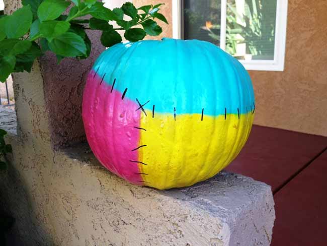 This playful pumpkin is painted with four different, bright colors—turquoise, pink, yellow, and red—separated by black stitches, giving it a fun, Frankenstein-inspired look.