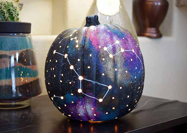 A dark pumpkin glows from within, painted with a stunning galaxy scene of deep blues, purples, and pinks, dotted with small lights that form a bright constellation.