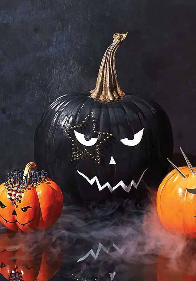 A large black pumpkin is decorated with jagged white eyes and a mouth, with a constellation-like star pattern over one eye created with metallic beads.