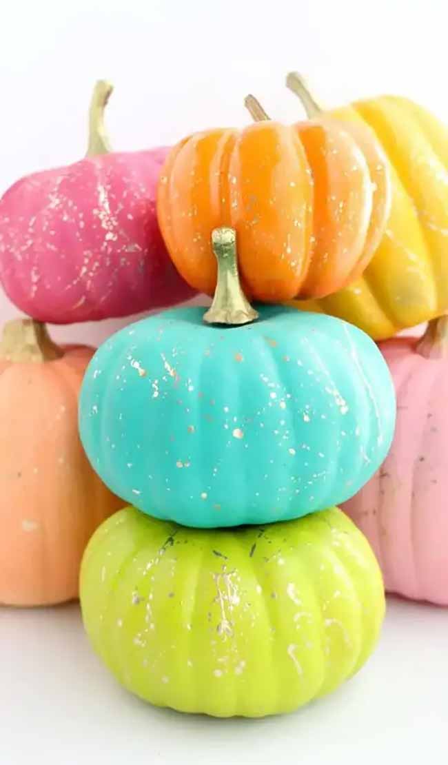 A cheerful pile of small pumpkins is painted in bright, candy-like colors such as teal, hot pink, and lime green, each with a light splatter of gold flakes.