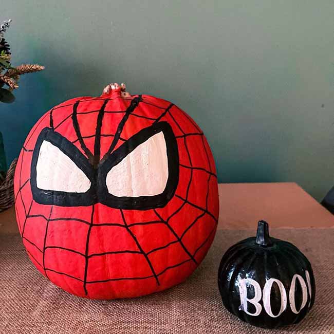 A bright red pumpkin is painted with a black spiderweb and large white eyes, creating a superhero-themed design next to a small black pumpkin labeled “BOO.”