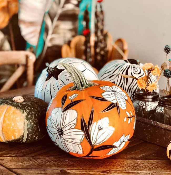 A large orange pumpkin is painted with white flowers, while pumpkins in the background feature black-and-white stripes and polka dots.