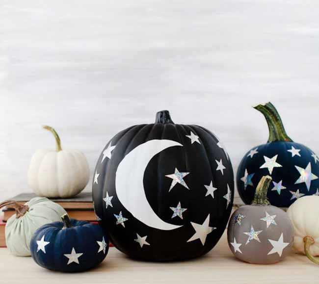 A large black pumpkin is decorated with a white crescent moon and glittery stars, accompanied by smaller navy and gray pumpkins with similar star patterns.